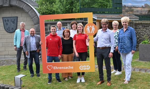 Fünf Männer und fünf Frauen lächeln freundlich und halten einen orangenen Rahmen mit der Aufschrift "Ehrensache".