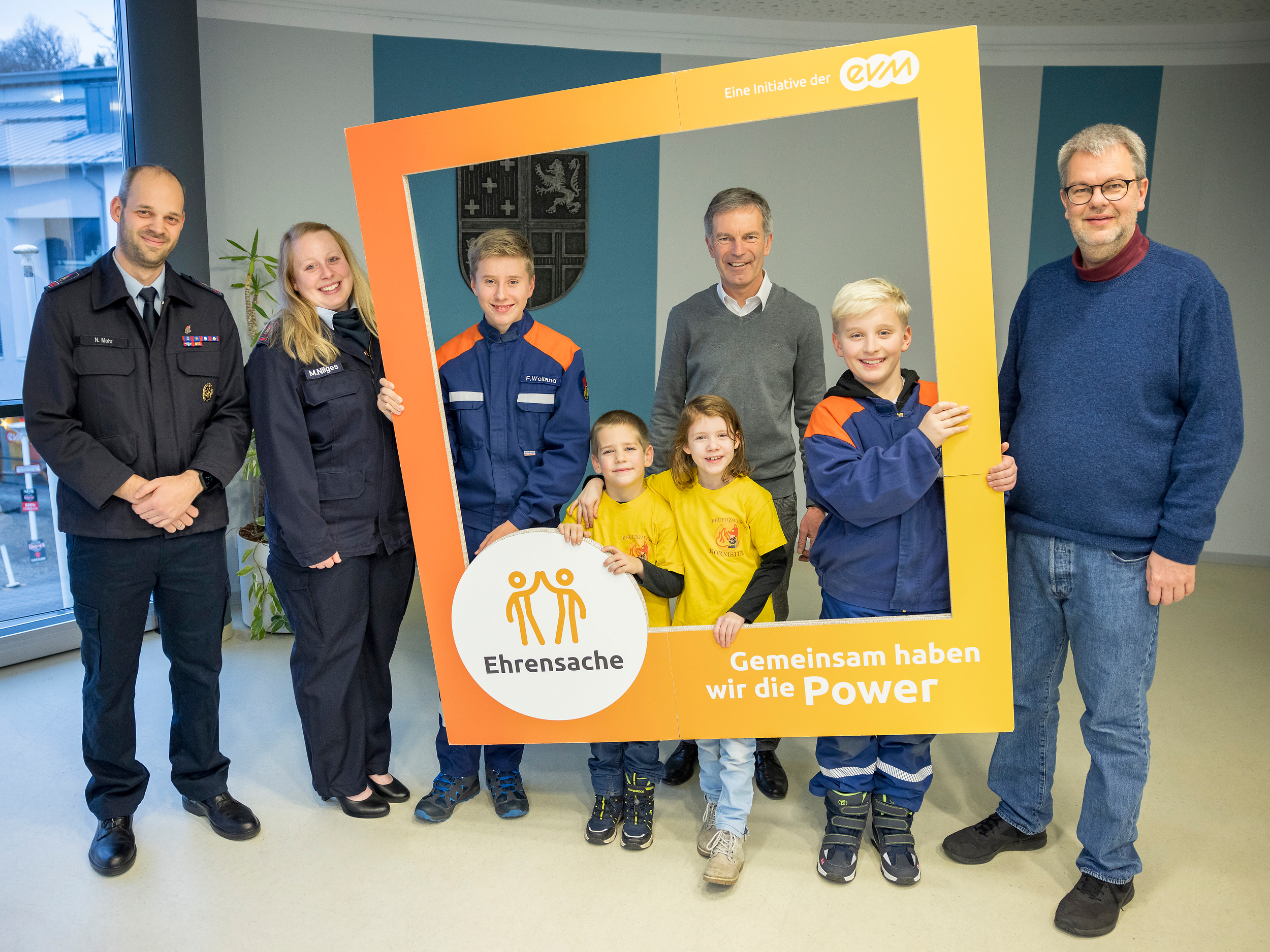 Vier Kinder und vier Erwachsene in Uniformen stehen in einem Raum und halten einen orangenen Rahmen mit der Aufschrift "evm Ehrensache". Sie lächeln freundlich.