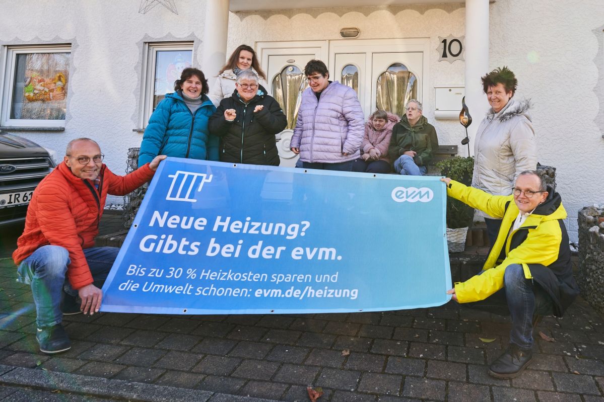 Eine Gruppe von Menschen hält ein blaues Banner vor einem Haus. Auf dem Banner steht: "Neue Heizung? Gibts bei der evm. Bis zu 30 % Heizkosten sparen und die Umwelt schonen: evm.de/heizung".