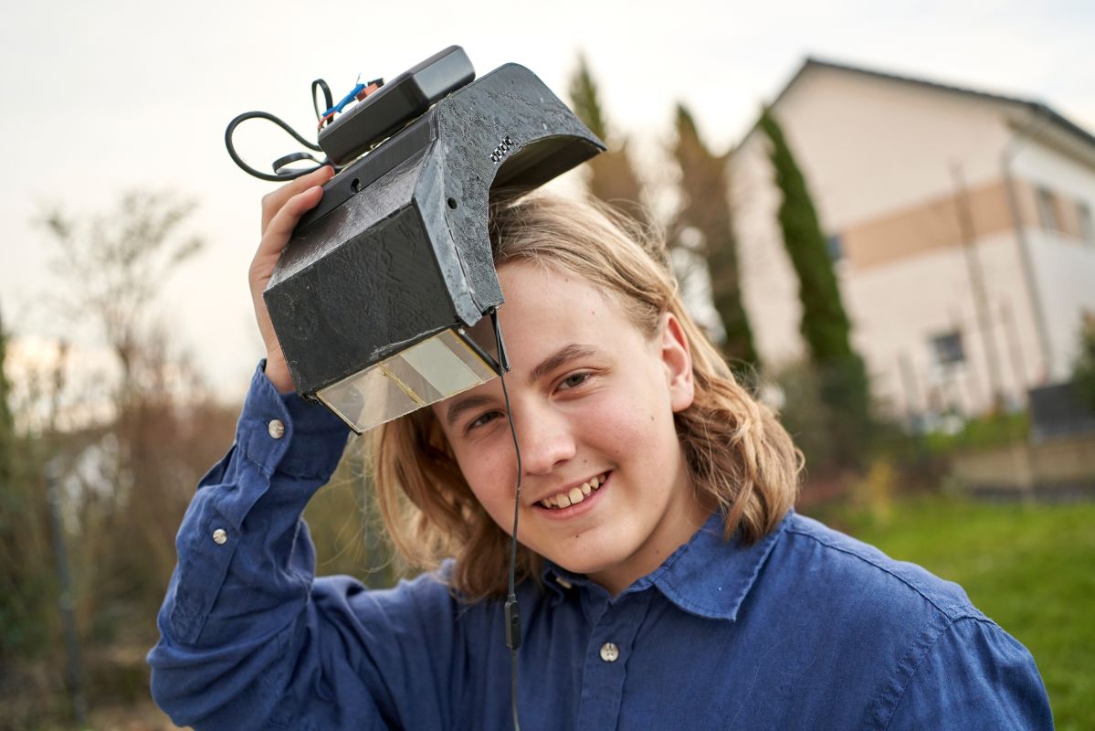 Ein Junge hält ein technisches Gerät über seinen Kopf und lächelt. Er trägt ein blaues Hemd. Im Hintergrund ist ein Haus und eine grüne Umgebung zu sehen.