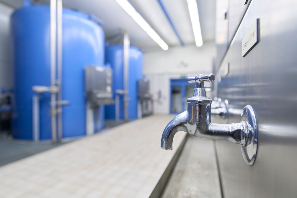 Ein Wasserhahn befindet sich an einer Wand mit Schildern; im Hintergrund sind große blaue Tanks in einem hell beleuchteten technischen Raum zu sehen.