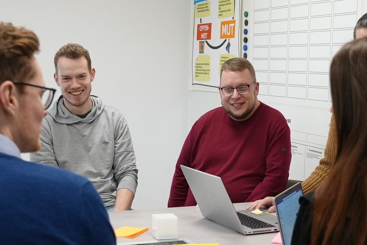 Mehrere Personen sitzen um einen Besprechungstisch und lächeln freundlich. auf dem Tisch stehen Computer.