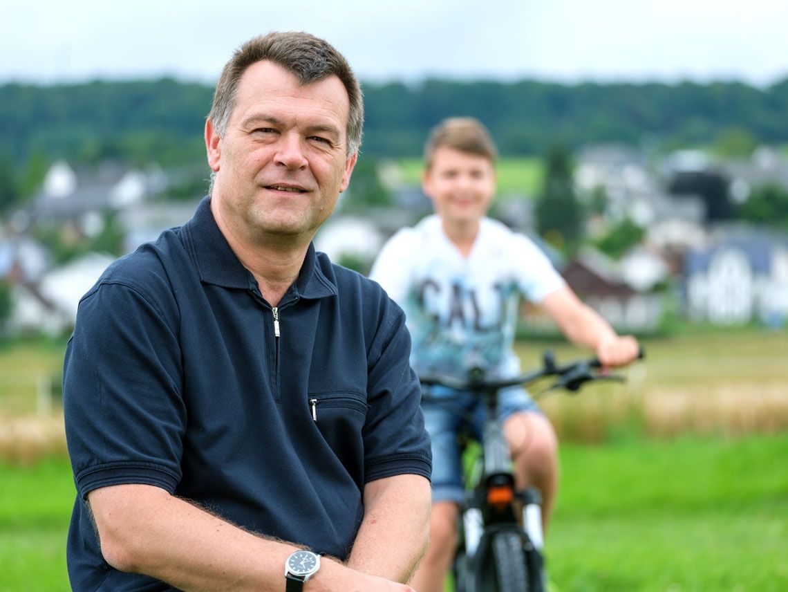 Ein lächelnder Mann sitzt auf einer Wiese, während im Hintergrund ein Junge auf einem Fahrrad fährt. Der Himmel ist bewölkt und es gibt unscharfe Häuser und Wälder.