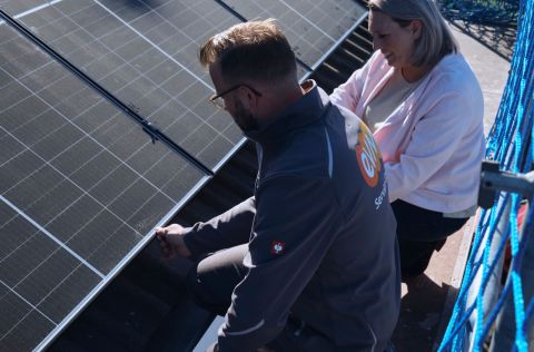Eine Person montiert Solarzellen auf einem Dach, begleitet von einer anderen Person, die zuschaut. Beide tragen Arbeitskleidung. Ein blaues Sicherheitsnetz umrahmt die Szene.
