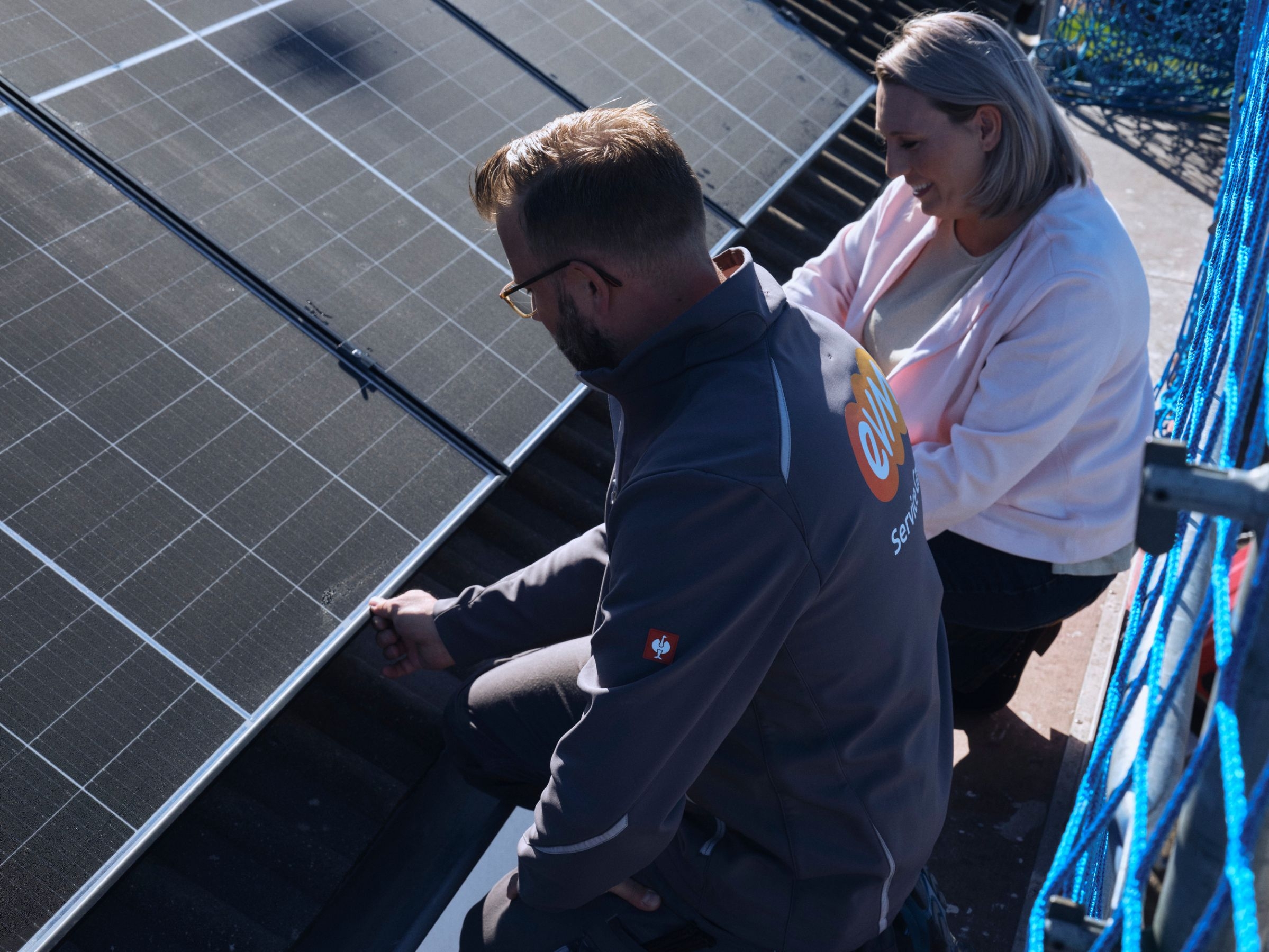Eine Person arbeitet an einer Solaranlage auf einem Dach, unterstützt von einer anderen Person. Beide tragen Arbeitskleidung. Die Umgebung wirkt industriell oder handwerklich.