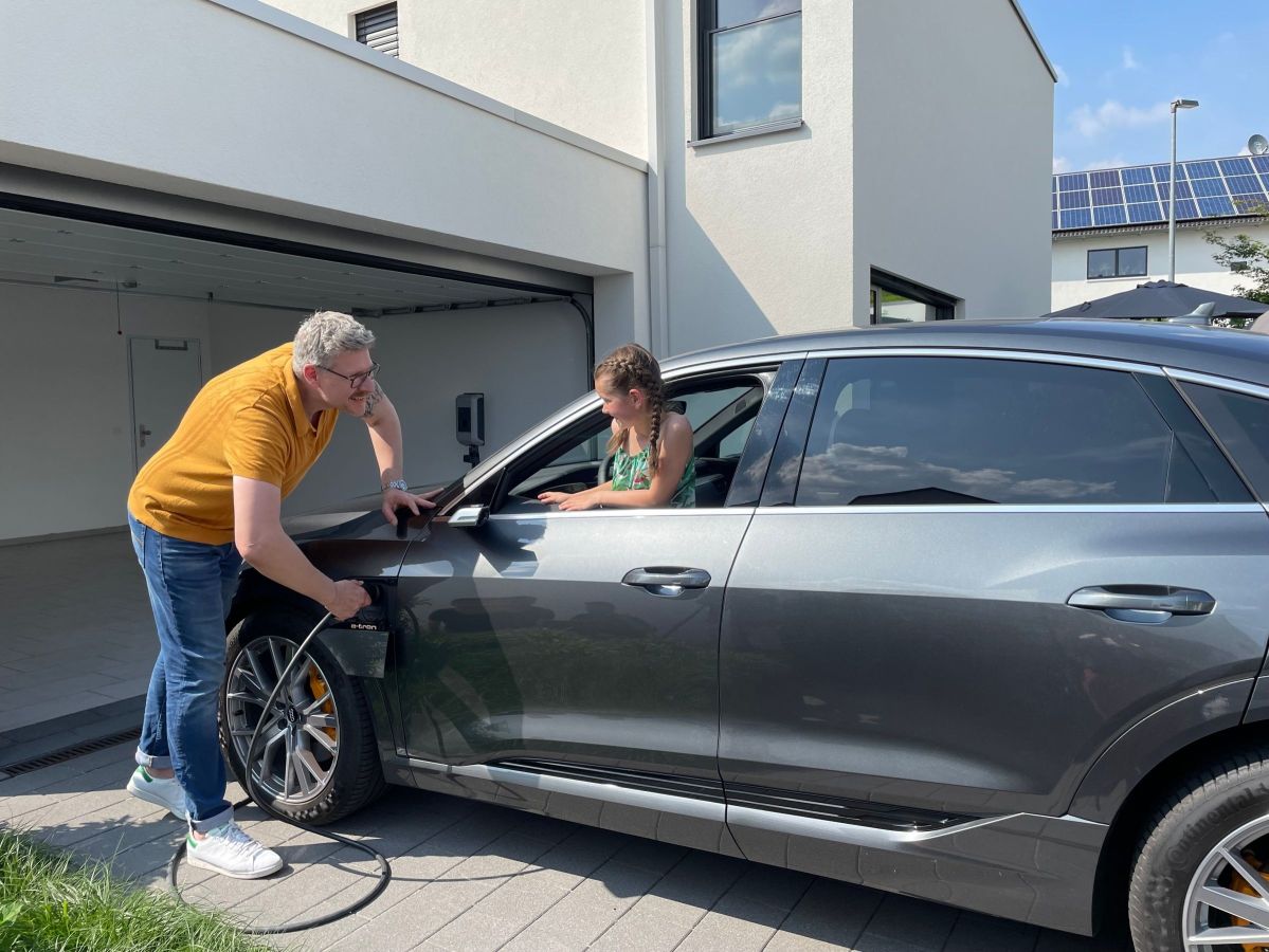 Ein Mann lädt ein Elektroauto in der Einfahrt eines modernen Hauses auf, während eine Frau im Auto sitzt. Im Hintergrund sind Solarpaneele auf dem Dach zu sehen.