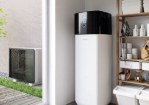 Ein großer weißer Heizkessel steht in einem Hauswirtschaftsraum neben einem offenen Regal mit Haushaltsgegenständen; eine Wärmepumpe ist draußen sichtbar neben einem hellen Gebäude und Rasen.