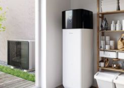 Ein großer weißer Heizkessel steht in einem Hauswirtschaftsraum neben einem offenen Regal mit Haushaltsgegenständen; eine Wärmepumpe ist draußen sichtbar neben einem hellen Gebäude und Rasen.