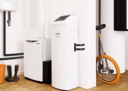 Ein weißes Heizsystem mit Display steht neben einem weiteren Gerät in einem modernen, hellen Raum mit Holzfußboden; daneben befinden sich ein orangefarbenes Einrad und Gummistiefel.