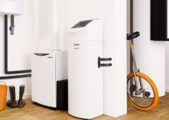 Ein weißes Heizsystem mit Display steht neben einem weiteren Gerät in einem modernen, hellen Raum mit Holzfußboden; daneben befinden sich ein orangefarbenes Einrad und Gummistiefel.