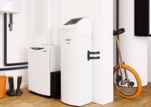 Ein weißes Heizsystem mit Display steht neben einem weiteren Gerät in einem modernen, hellen Raum mit Holzfußboden; daneben befinden sich ein orangefarbenes Einrad und Gummistiefel.