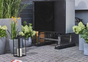 Ein großes, schwarzes Außengerät steht auf Metallstreben auf einem gepflasterten Boden. Daneben befindet sich ein Topf mit grünen Pflanzen. Im Hintergrund sind Holzwände erkennbar.