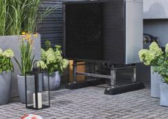 Ein großes, schwarzes Außengerät steht auf Metallstreben auf einem gepflasterten Boden. Daneben befindet sich ein Topf mit grünen Pflanzen. Im Hintergrund sind Holzwände erkennbar.