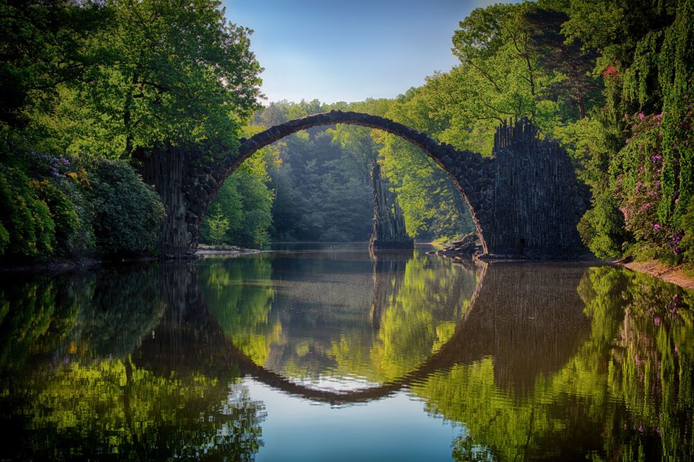 Eine steinerne Bogenbrücke spannt sich über einen ruhigen Fluss, reflektiert im Wasser, umgeben von üppigen, grünen Bäumen und dichtem Wald unter klarem, blauem Himmel.