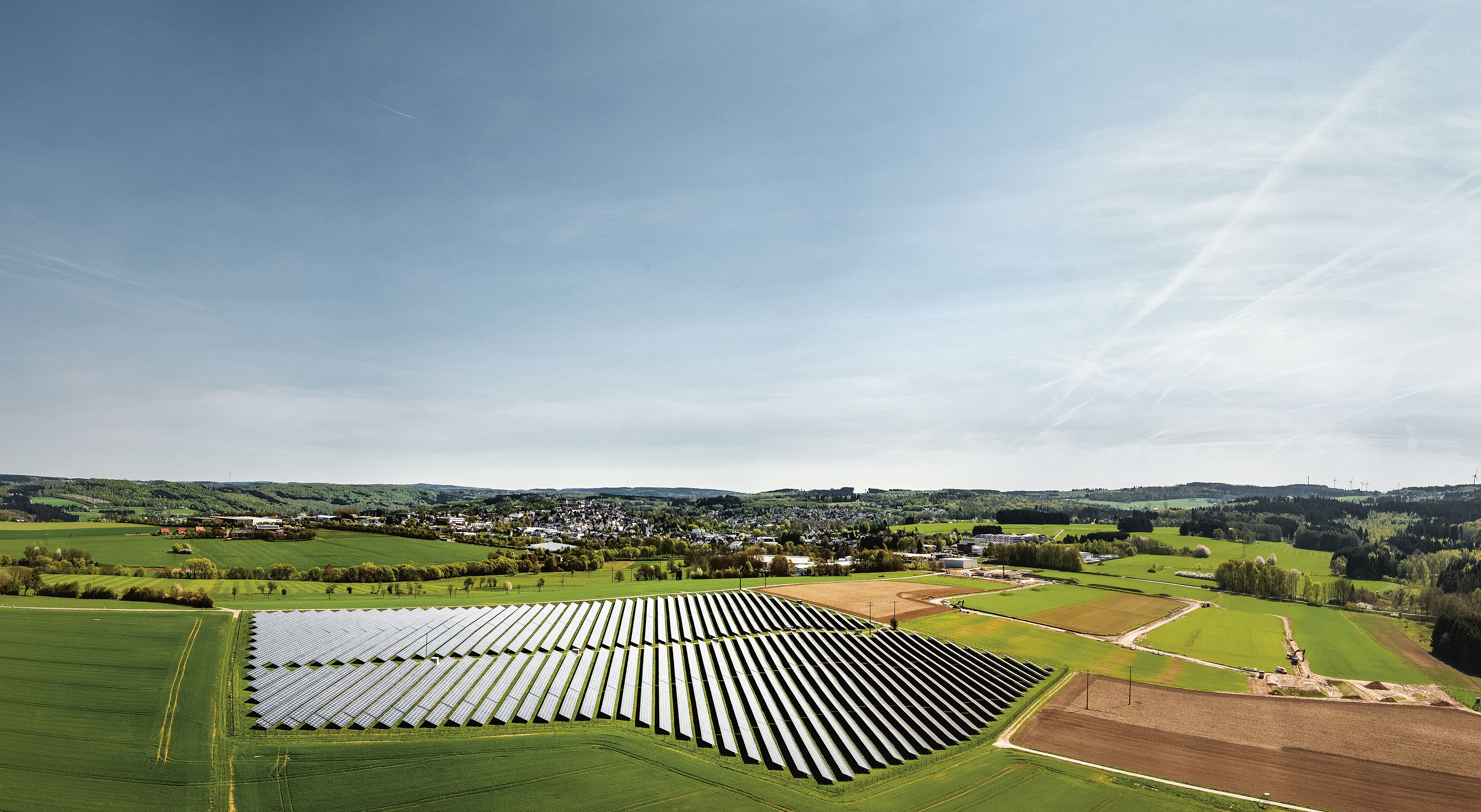Eine große Solaranlage liegt inmitten grüner Felder, umgeben von sanften Hügeln und einer kleinen Stadt im Hintergrund unter klarem Himmel.