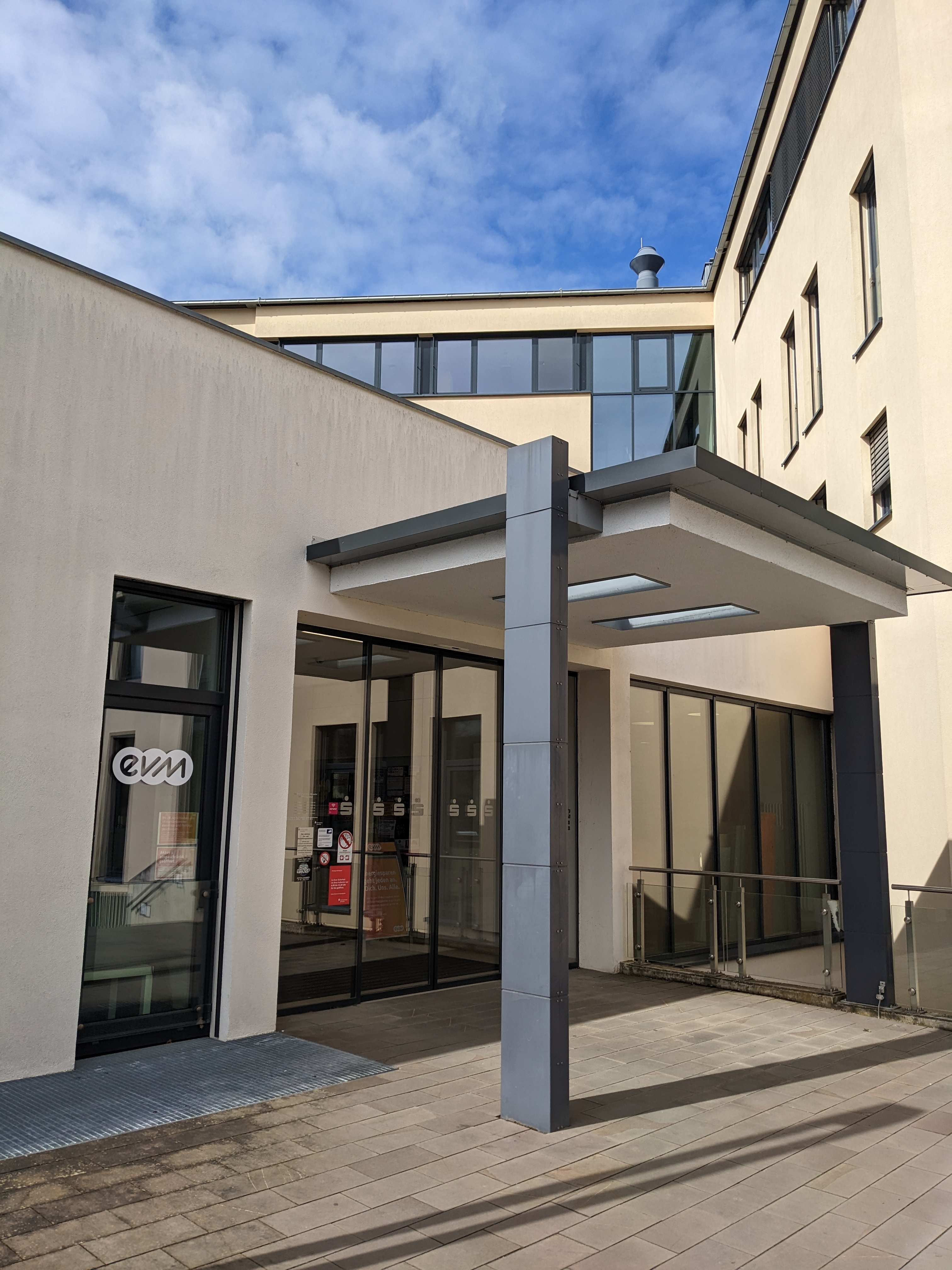 Ein modernes Eingangsportal mit Glasdach und Säulen führt in ein Gebäude. Im Hintergrund sind Fenster zu sehen. An einer Scheibe klebt ein Logo mit der Aufschrift "evm".