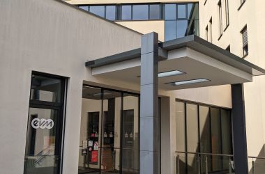 Ein modernes Eingangsportal mit Glasdach und Säulen führt in ein Gebäude. Im Hintergrund sind Fenster zu sehen. An einer Scheibe klebt ein Logo mit der Aufschrift "evm".