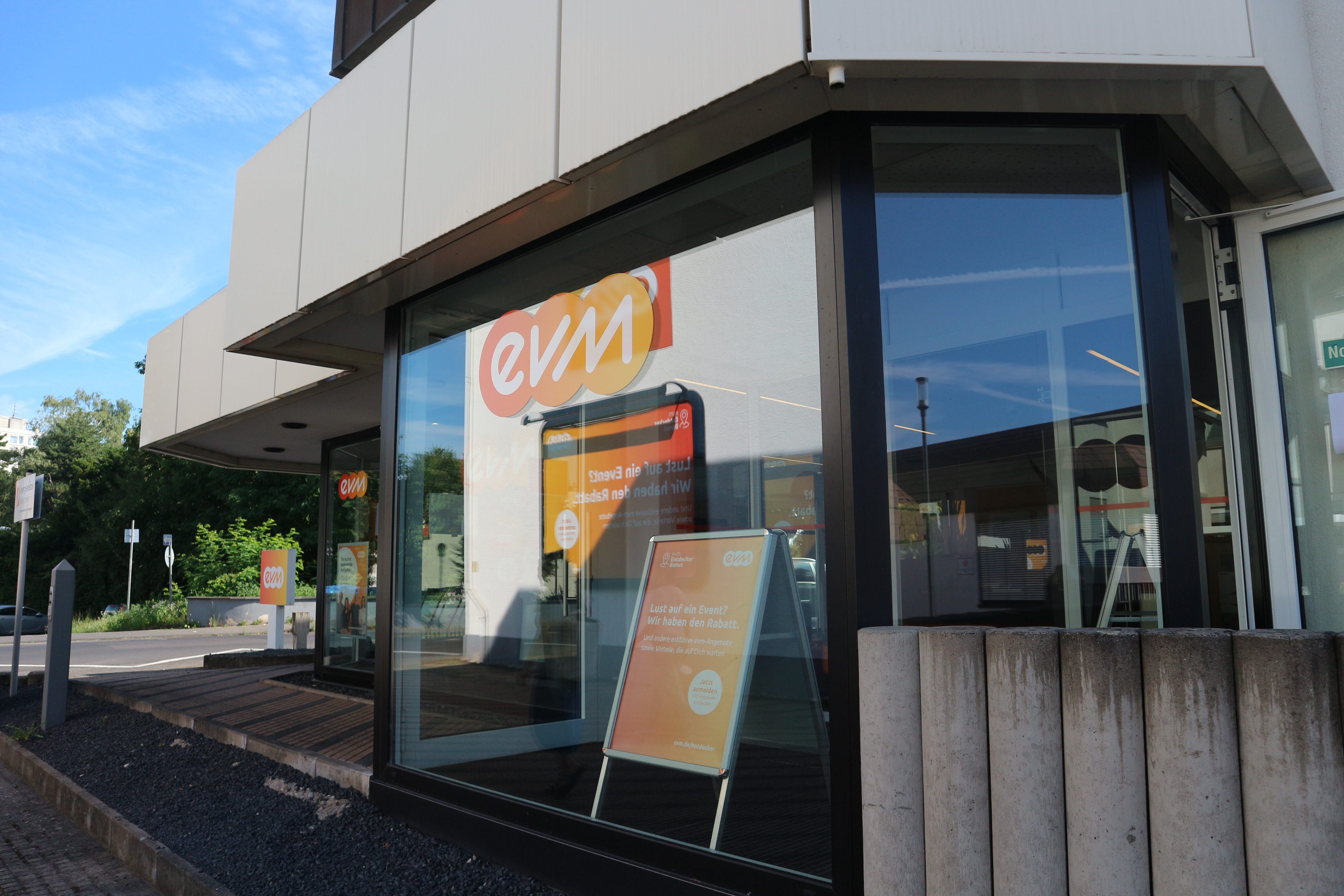 Der Schaukasten zeigt ein Plakat von "evm" in einem modernen, verglasten Gebäudeeingang. Weitere Plakate und Hintergrundlandschaft spiegeln sich in den Fenstern wider.
