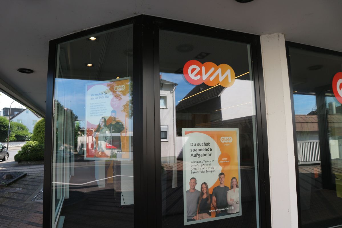 Eine Glastür mit evm-Logo trägt ein Plakat: "Du suchst spannende Aufgaben? Komm ins Team der evm-Gruppe... Jetzt bewerben!" Innerhalb einer städtischen Umgebung mit Bürgersteig.