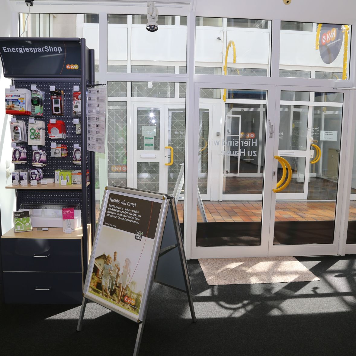 Ein Regal mit Energiesparprodukten steht neben einer Glastür in einem hellen Innenraum.