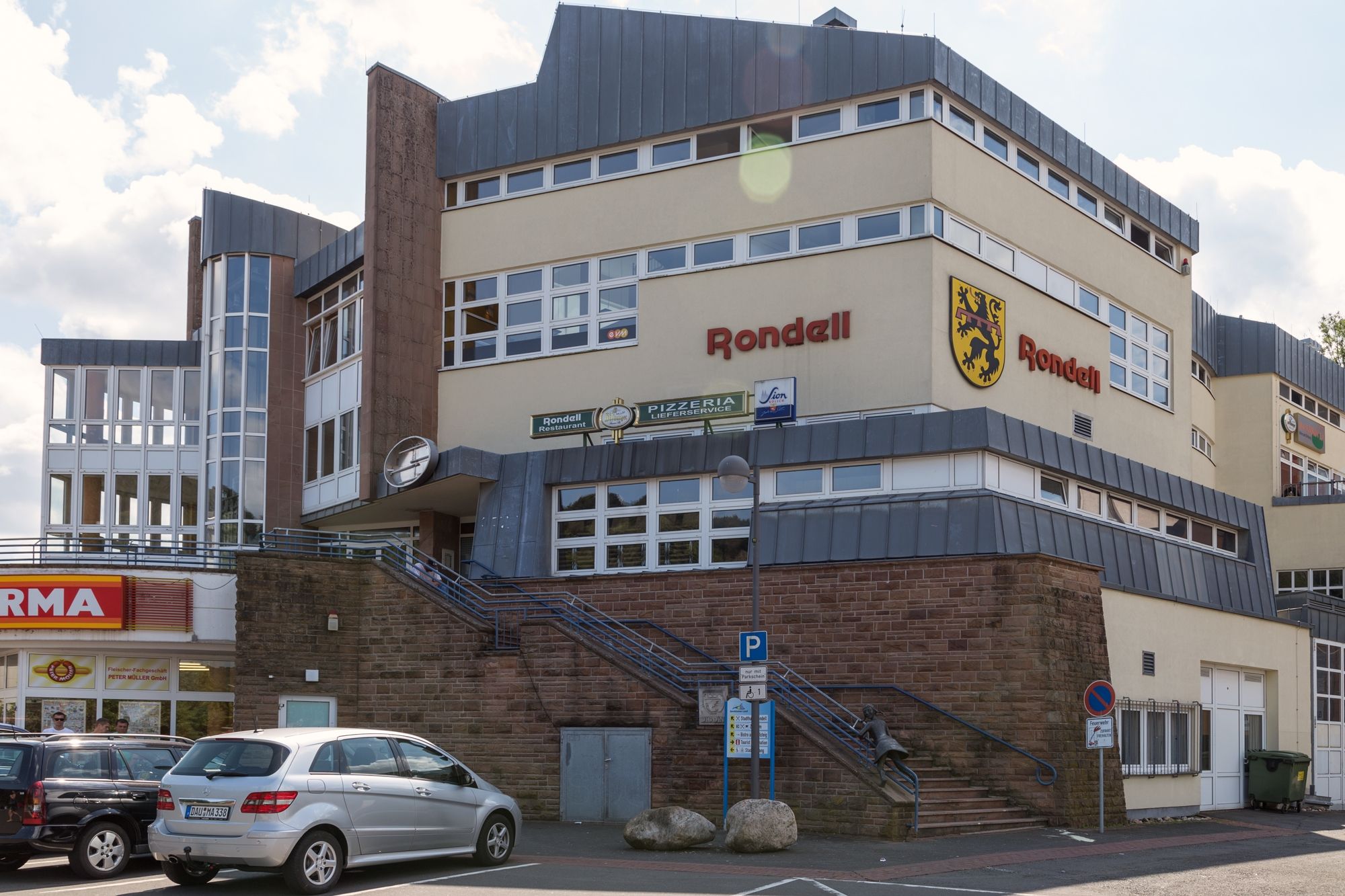 evm-Kundenzentrum Gerolstein, Ansicht von außen. Ein beige-gelbes Gebäude mit dem Schriftzug "Rondell" und einem Löwenwappen, Treppen führen hinauf, umgeben von Straßenverkehrsschildern und einem "NORMA"-Laden im Vordergrund.