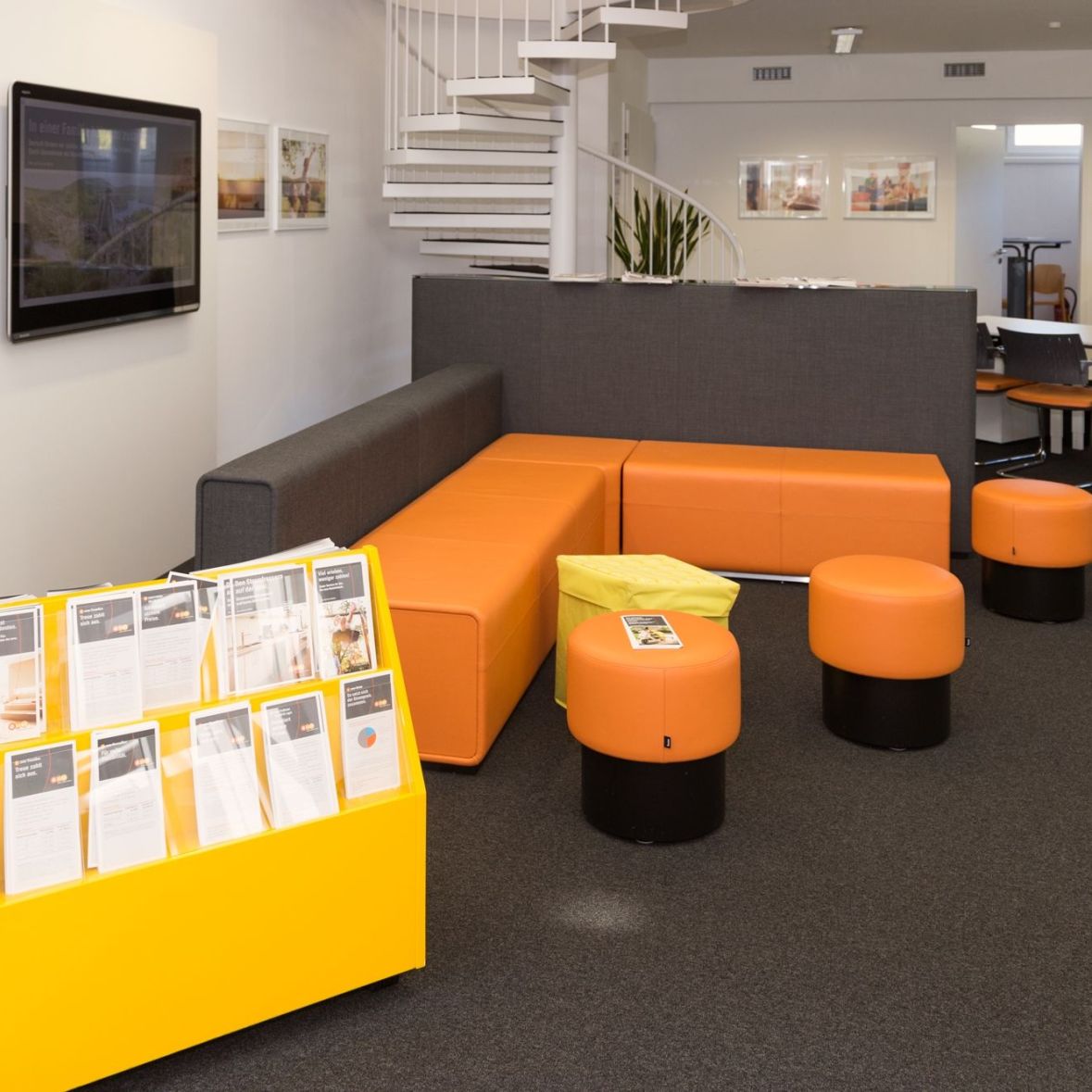 Ein Wartebereich mit einer grauen und orangen Sitzgruppe und einem gelben Prospektständer. Im Hintergrund befinden sich Bürotische, Stühle, eine Wendeltreppe und Bilder an der Wand.