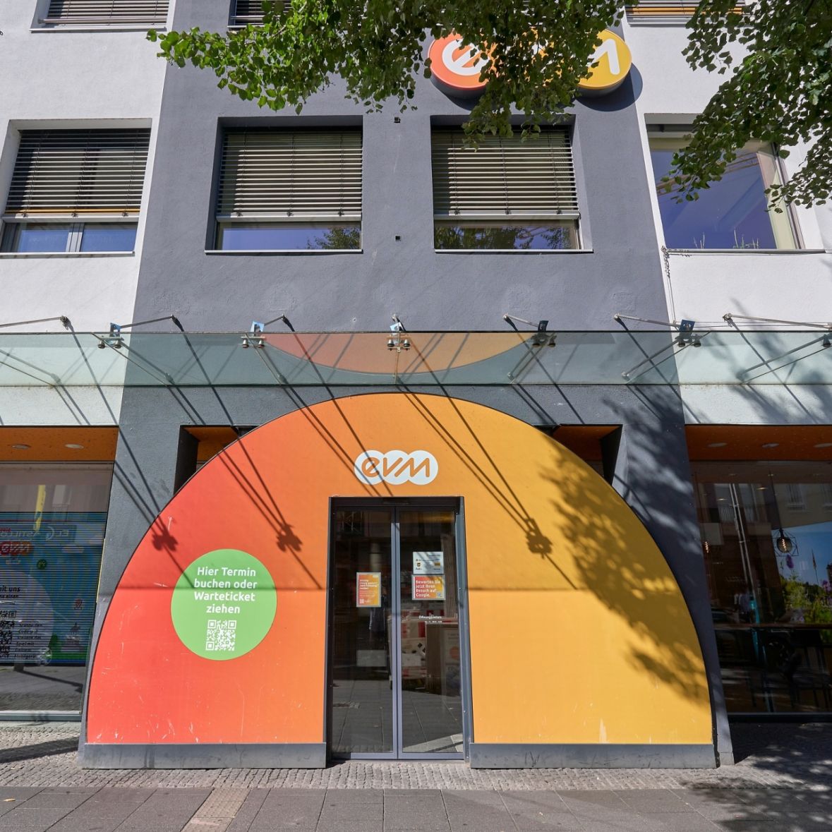 Eine halbkreisförmige orange-rote Wand mit einer Glastür in der Mitte und evm-Logo, vor einem Geschäft in einer Fußgängerzone.