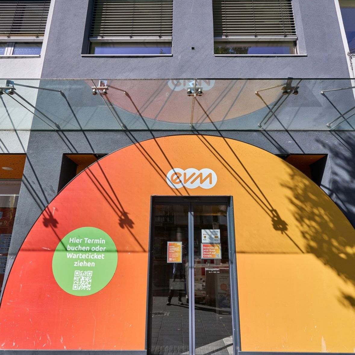 Eine halbkreisförmige orange-rote Wand mit einer Glastür in der Mitte und evm-Logo, vor einem Geschäft in einer Fußgängerzone.