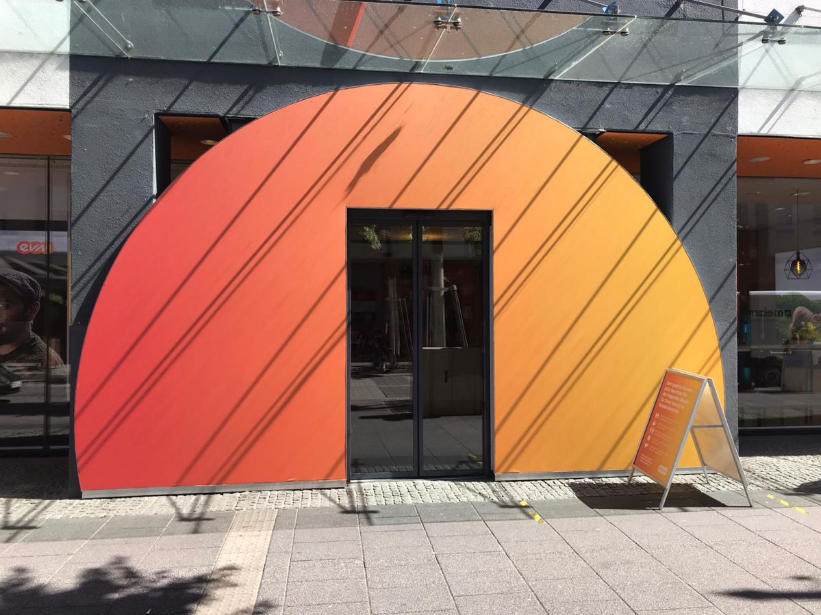 Eine halbkreisförmige orange-rote Wand mit einer Glastür in der Mitte, vor einem Geschäft in einer Fußgängerzone. Neben der Tür steht ein beschriftetes Schild.