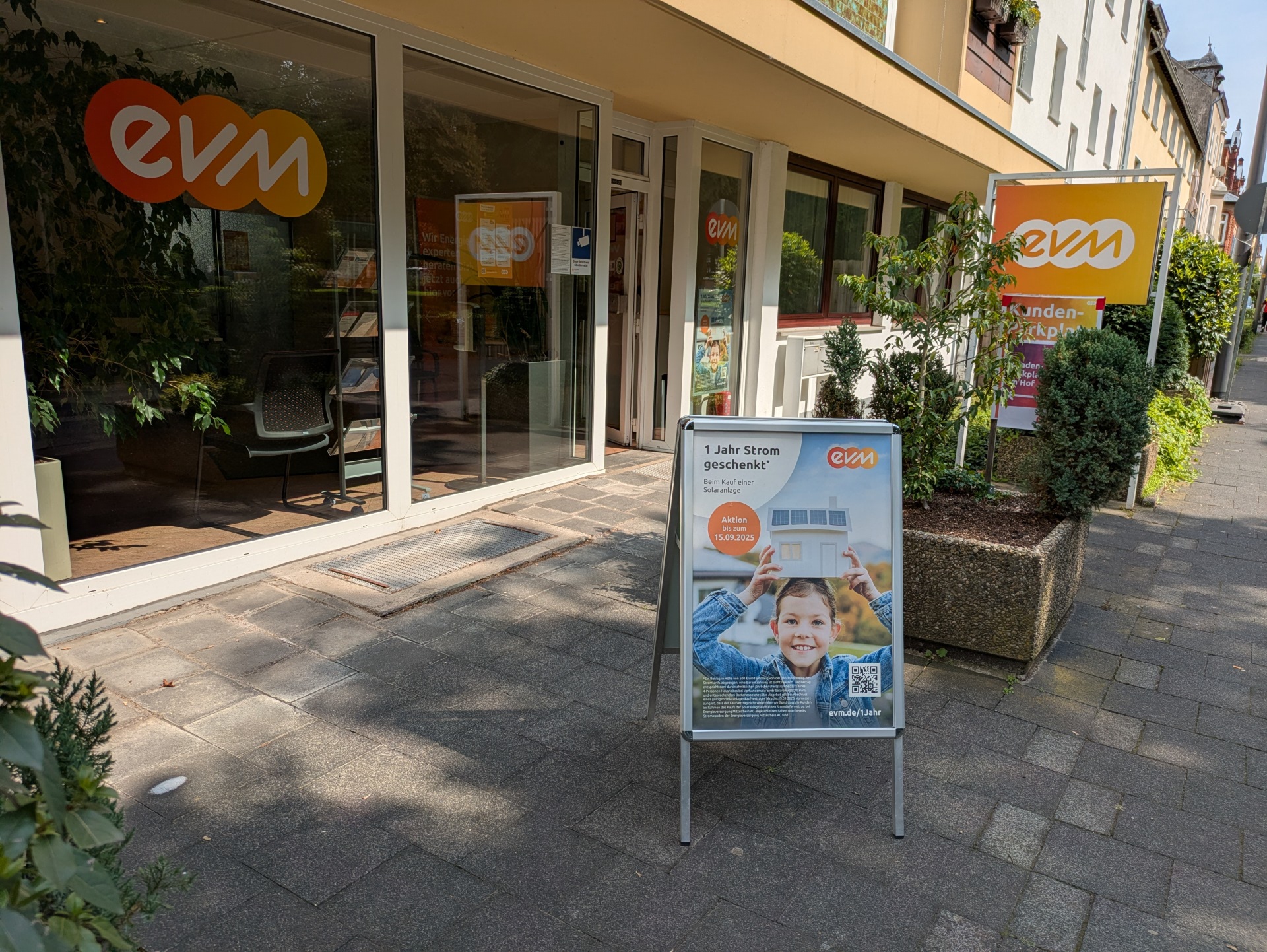 Ein Geschäft mit gläserner Front zeigt das Logo „evm“. Davor stehen Pflanzkübel und ein Werbeplakat auf einem Gehweg.