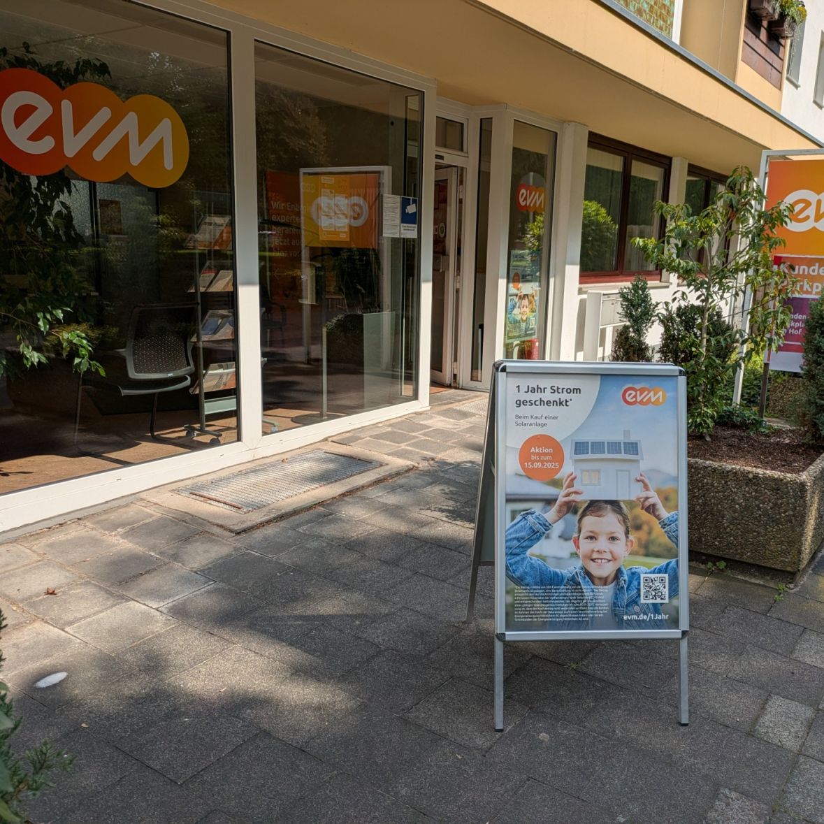 Ein Geschäft mit gläserner Front zeigt das Logo „evm“. Davor stehen Pflanzkübel und ein Werbeplakat auf einem Gehweg.