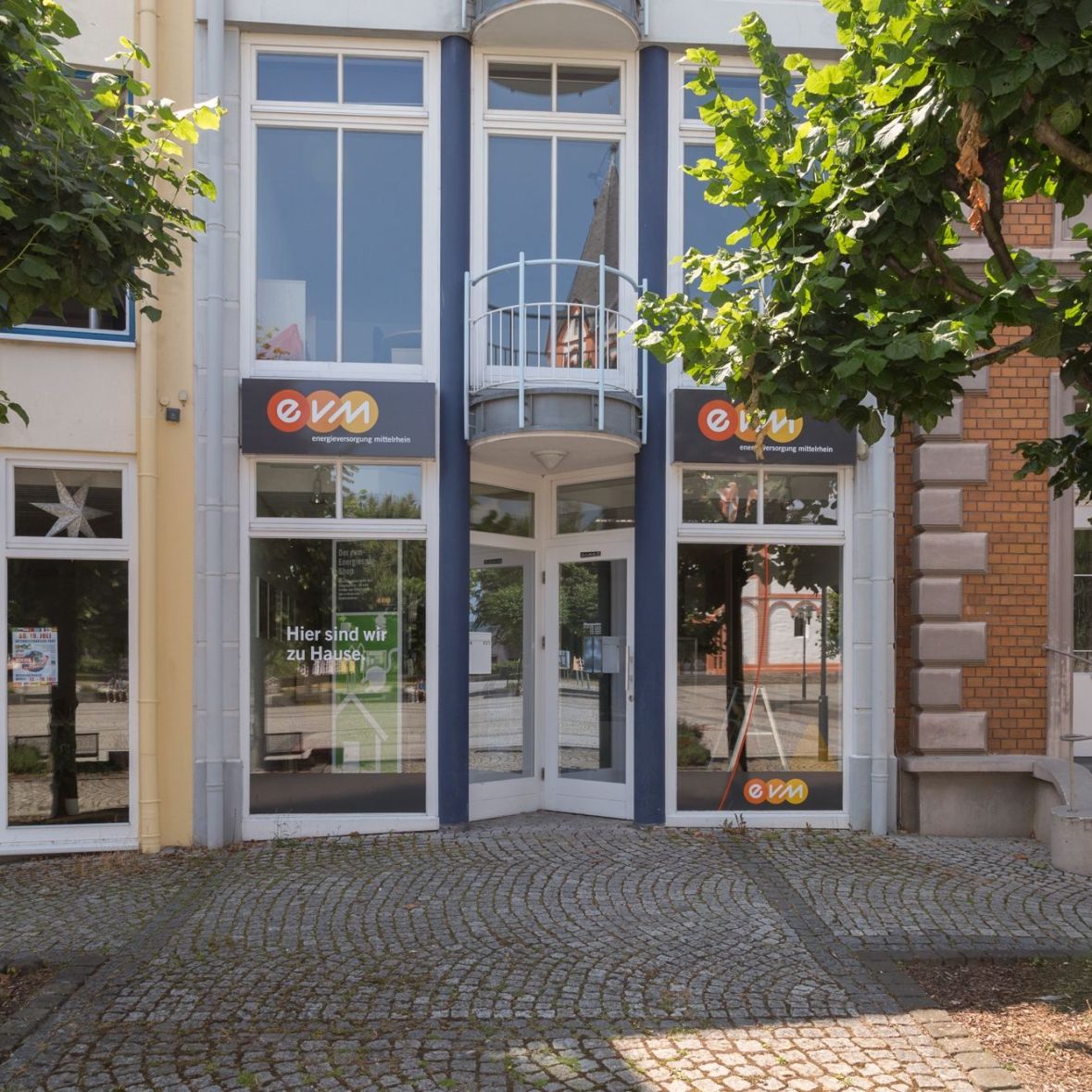 Ein modernes Gebäude mit großen Fenstern zeigt zwei Schilder mit "evm". Bäume stehen davor, und der Gehweg aus Kopfsteinpflaster verläuft entlang der Fassade.