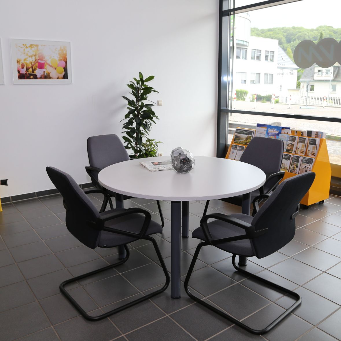 Ein runder Tisch mit vier dunklen Stühlen steht in einem modernen Raum. Eine Pflanze und ein Bild an der Wand; Fenster zeigen städtische Umgebung.