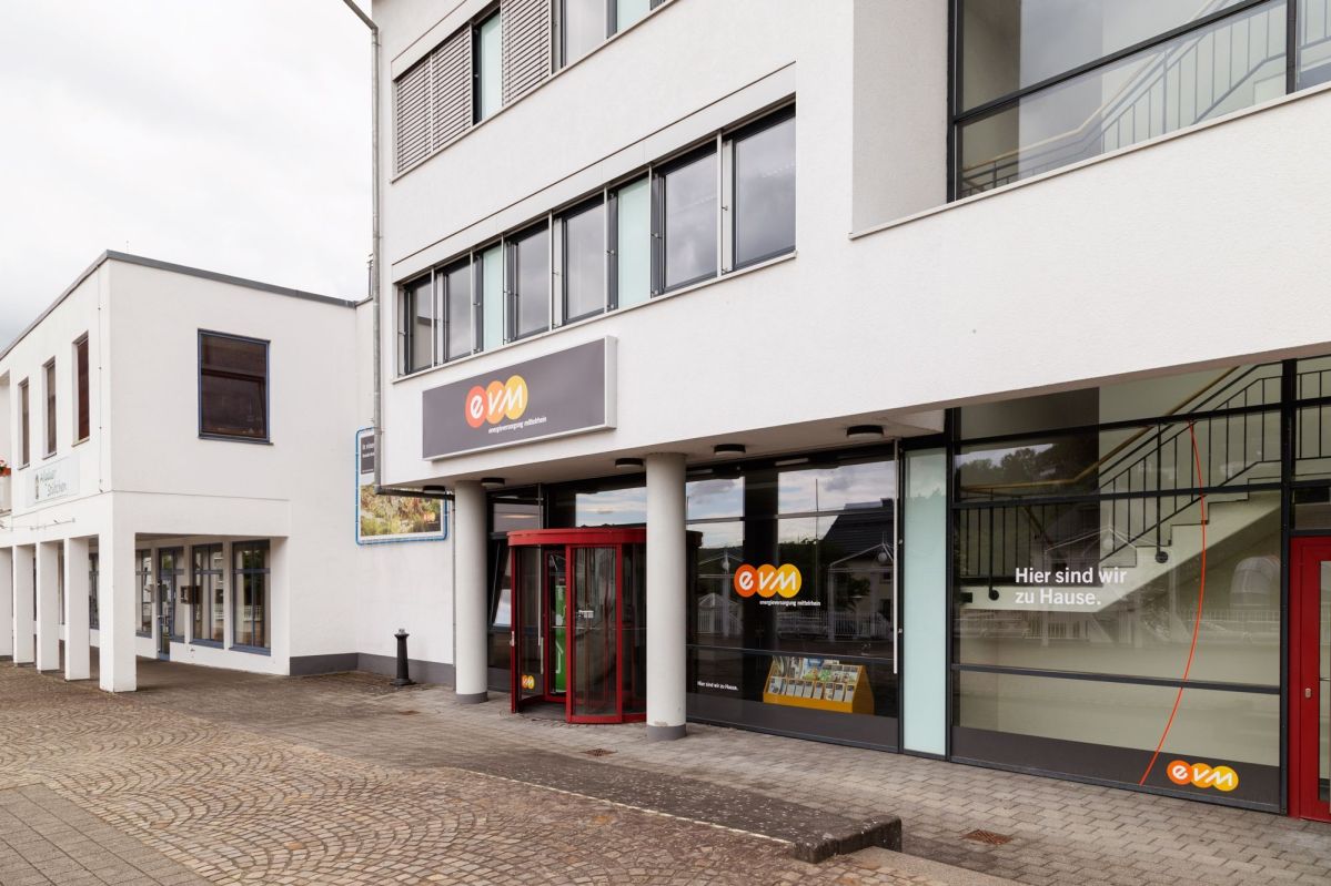 Ein Bürogebäude mit einer gewölbten Glasfront, der Eingang hat ein rotes Drehkreuz. Über dem Eingang ist ein Schild mit dem Logo "evm". Text: "Hier sind wir zu Hause."
