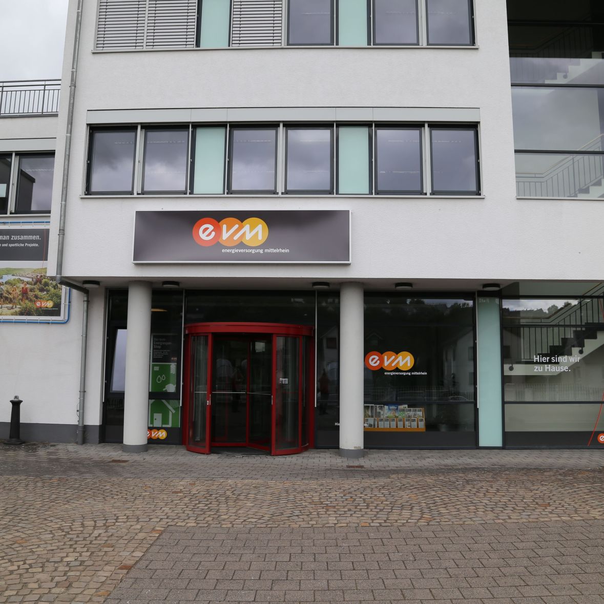Ein modernes Gebäude mit Glasfassade. Ein roter Eingang dreht sich. Auf dem Schild steht „evm“. Steinpflaster davor.