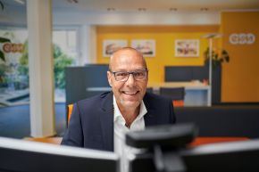 evm Kundenzentrum Eine lächelnde Person sitzt an einem Schreibtisch in einem modernen Büro. Im Hintergrund sind orangefarbene Wände, ein Tisch, Pflanzen und Dekorationen zu sehen. Büroatmosphäre vermittelt Professionalität.