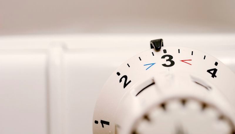 Ein weißer Heizkörper-Thermostat zeigt die Zahl 3 an. Blaue und rote Markierungen deuten Kälte und Wärme an. Der Hintergrund ist weiß und unscharf, möglicherweise eine Wand.