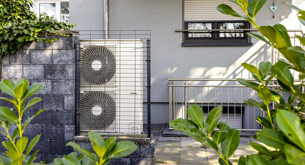 Das Außenelement einer Wärmepumpe an einer Hauswand. Drumherum stehen etliche Pflanzen