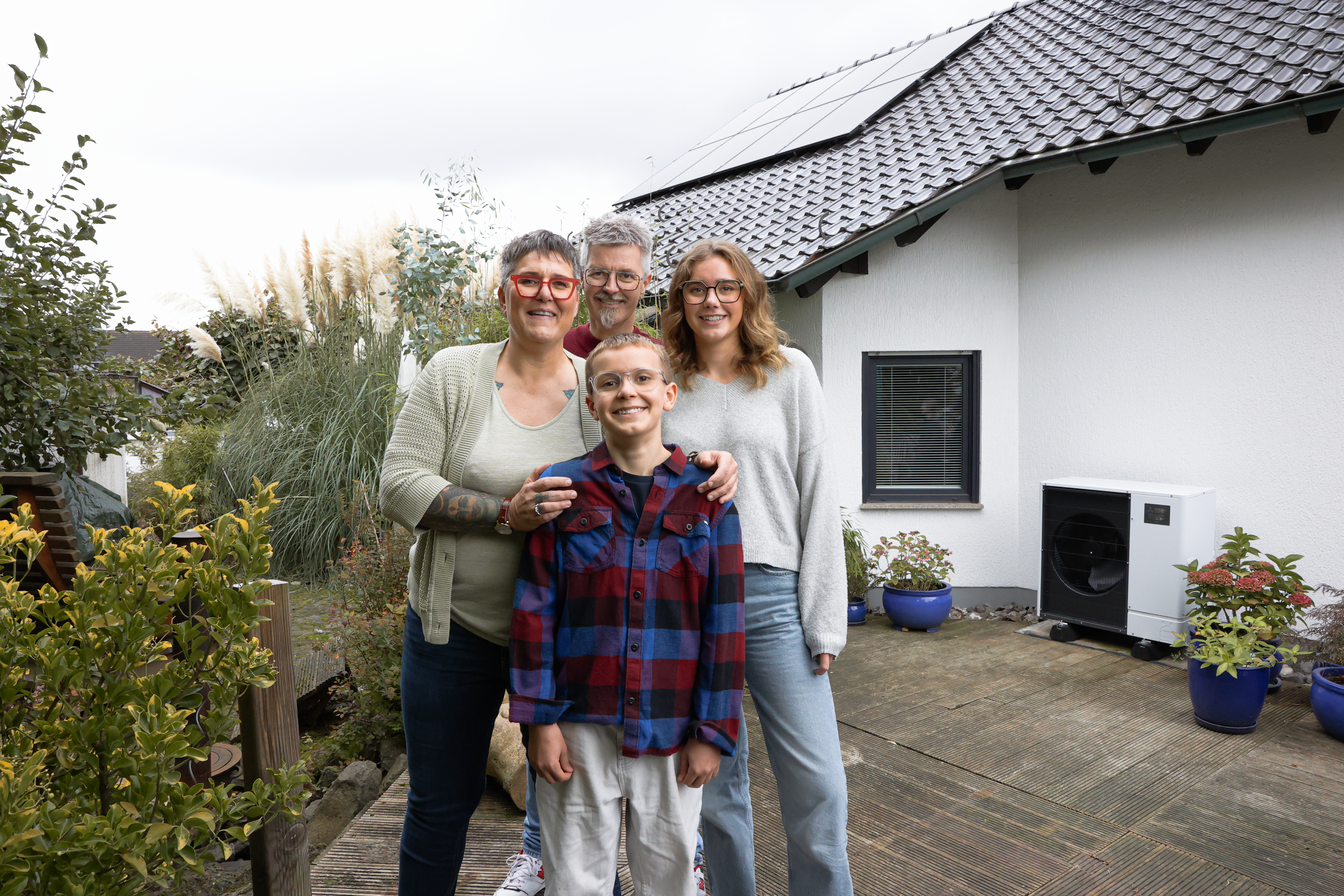 Eine perfekte Kombination: Wärmepumpe und PV-Anlage Eine vierköpfige Familie steht lächelnd vor einem modernen Haus mit Solarpanelen auf dem Dach. Sie befinden sich im Garten, umgeben von Pflanzen und Blumen in Töpfen.