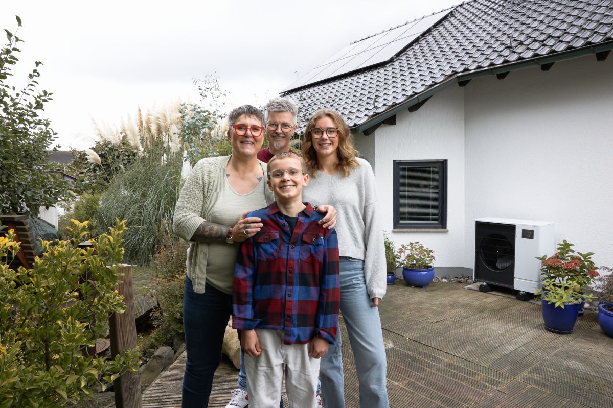 Eine perfekte Kombination: Wärmepumpe und PV-Anlage Eine vierköpfige Familie steht lächelnd vor einem modernen Haus mit Solarpanelen auf dem Dach. Sie befinden sich im Garten, umgeben von Pflanzen und Blumen in Töpfen.