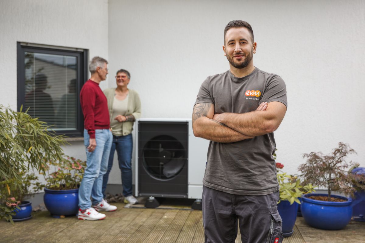 Das Foto zeigt eine Szene im Außenbereich vor einer weißen Hauswand. Im Vordergrund steht eine Person mit verschränkten Armen, bekleidet mit einem grauen T-Shirt, auf dem ein Logo mit der Aufschrift „evm“ und „wir sind da“ zu sehen ist. Die Person trägt dunkle Arbeitshose und steht auf einer Holzterrasse. Im Hintergrund befindet sich eine große Wärmepumpe, die direkt an der Hauswand installiert ist. Links daneben stehen zwei weitere Personen, die sich miteinander unterhalten.
