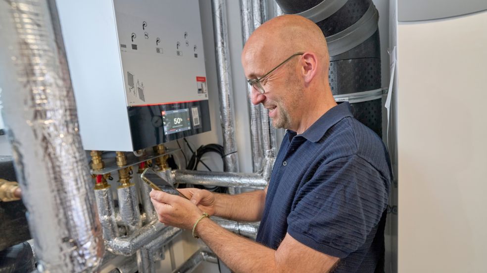 Thomas Schmitz steht vor einer modernen Heizungsanlage mit sichtbaren Rohrleitungen und einem Steuergerät, das 50 °C anzeigt. Er bedient die Anlage per Smartphone.