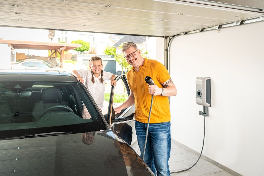 Ein Mann in einem gelben Poloshirt und seine Tochter stehen an einem Auto. Er hält ein e-Ladekabel