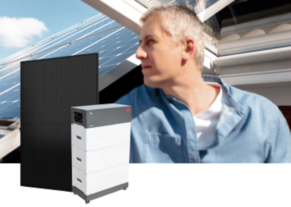 Ein Mann lächelt zufrieden. Neben ihm steht eine Solarbatterie vor einem Haus mit Solarmodulen auf dem Dach.