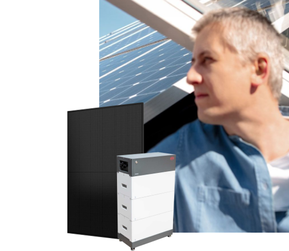 Ein Mann lächelt zufrieden. Neben ihm steht eine Solarbatterie vor einem Haus mit Solarmodulen auf dem Dach.