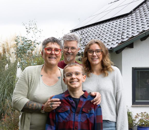Eine Familie steht vor einem Haus mit PV-Anlage und lächelt freundlich