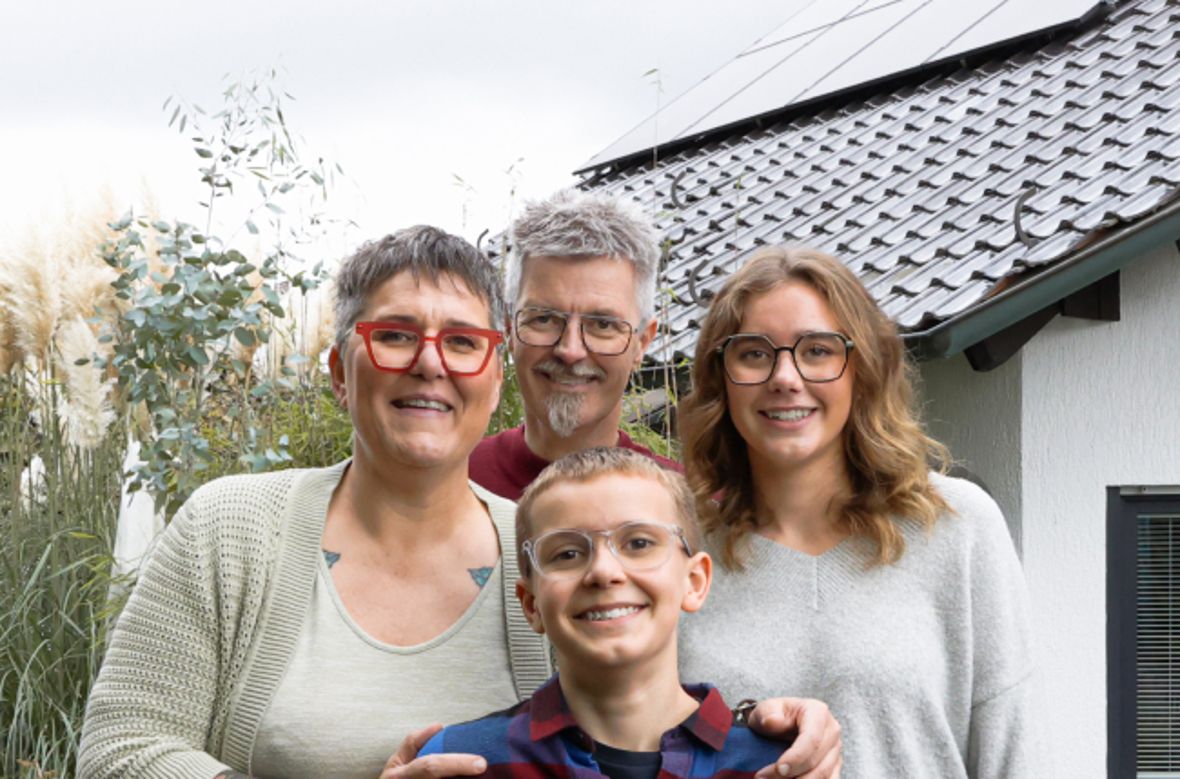 Eine Familie steht vor einem Haus mit PV-Anlage und lächelt freundlich