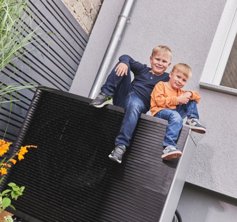 Zwei Kinder sitzen auf einer Wärmepumpe in einem Garten