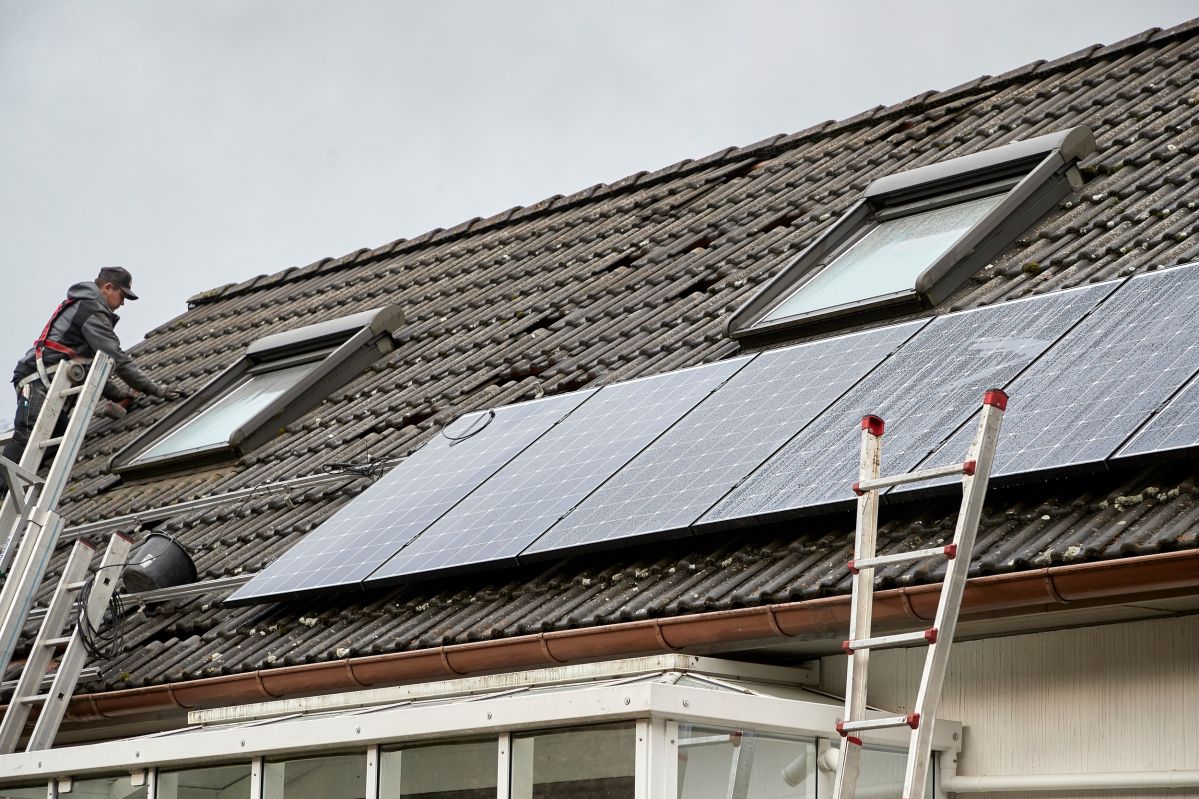 Ein Arbeiter an einer Leiter installiert Solarpaneele auf einem geneigten Ziegeldach eines Hauses mit zwei Dachfenstern. Zwei Leitern sind an verschiedenen Stellen des Daches platziert.