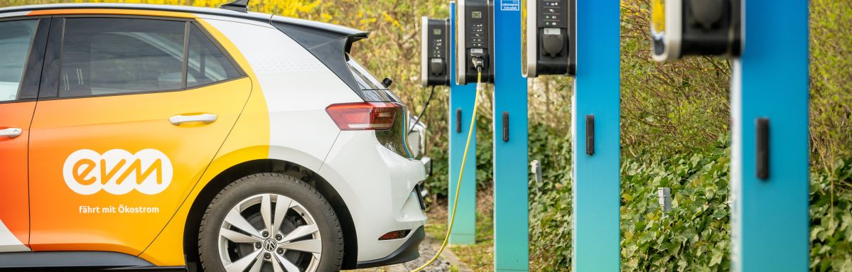 Ein weißes Elektroauto wird an eine von drei blauen Ladestation angeschlossen. Im Hintergrund sind Büsche zu sehen. Die Ladestationen sind durch ein gelbes Kabel mit dem Auto verbunden.